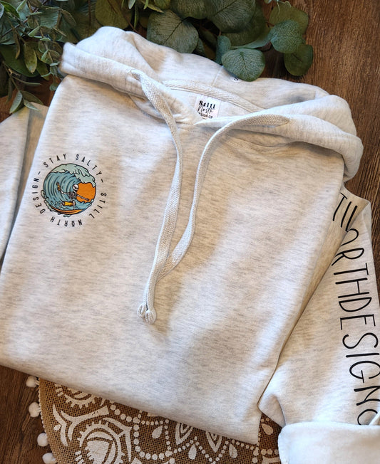 Beige hoodie with a logo and text on a wooden surface with greenery in the background